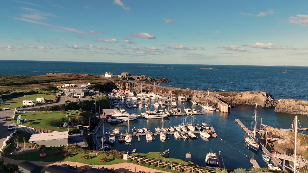 Slow forward flight over Beaucette Marina Guernsey in bright sunshine with full view of marina and berthed boats and entrance with blue sky. Fort Doyle in the background