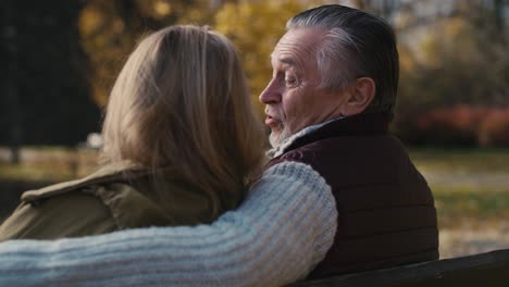 vista trasera pareja de ancianos caucásicos sentados en el banco en el parque y abrazándose