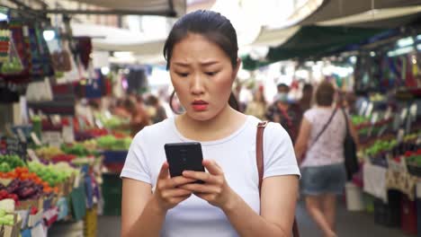 a woman feels concerned while browsing her smartphone in a busy market, surrounded by vibrant fruits and vegetables, capturing a moment of urban life and emotion.