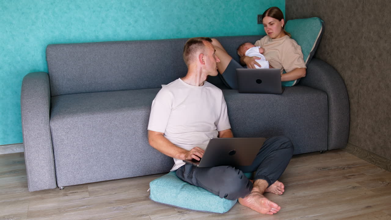 Working mom breastfeeding a baby. Male sitting at the sofa uses laptop and talks to his wife.