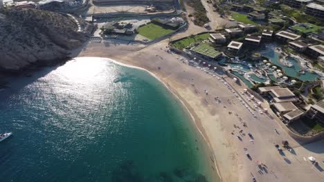 Aerial-parallax-shot-of-the-wonderful-medano-beach-in-cabo-san-lucas,-mexico-overlooking-the-blue-sea-with-boats-in-the-water,-hotel-buildings-and-majestic-mountains-in-the-background-on-a-sunny-day