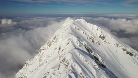 Snow-capped-Piatra-Craiului-mountain-peak-above-clouds,-sunlit-with-clear-blue-sky,-aerial-view