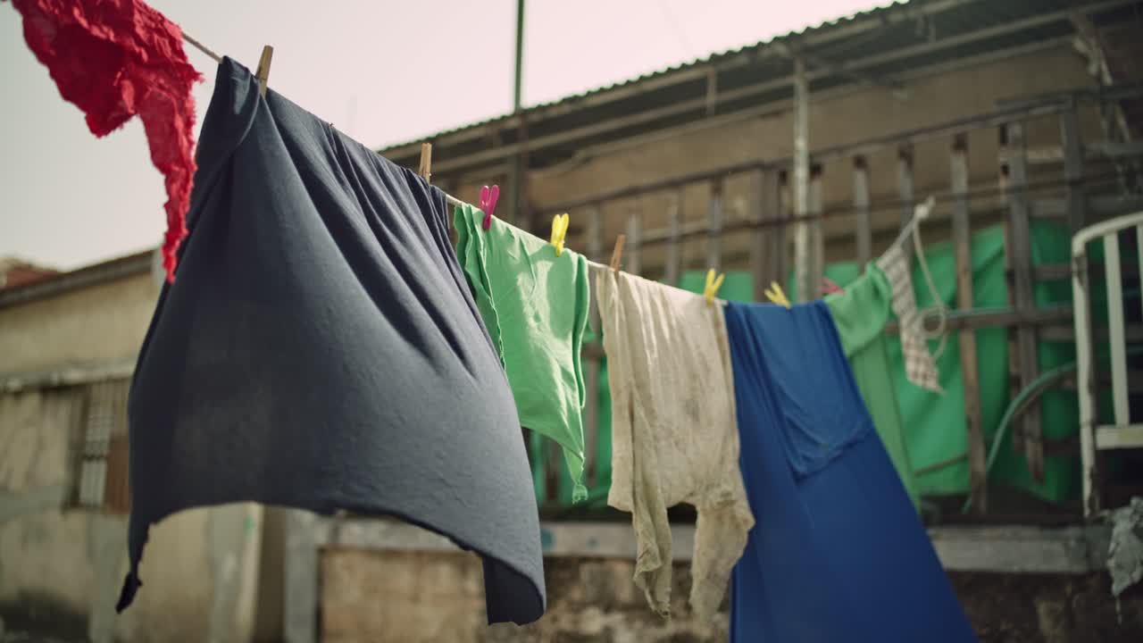 Old rugged dirty clothes and fabrics hanging from laundry line drying in poor neighborhood or street alley in summer or hot day. Shabby low income simple living. Windy day outdoors in the slums