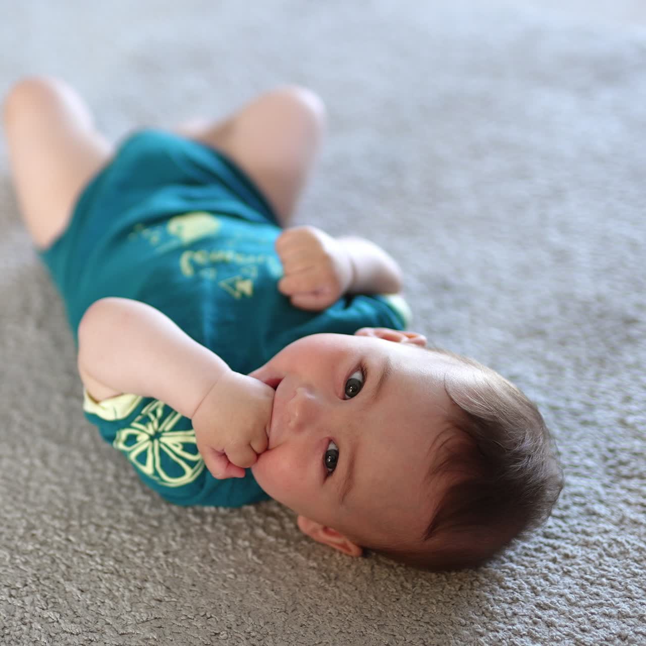 Lovely kid lies on the floor his head to the camera. Baby chewing his fist and smiling sweetly. Blurred backdrop