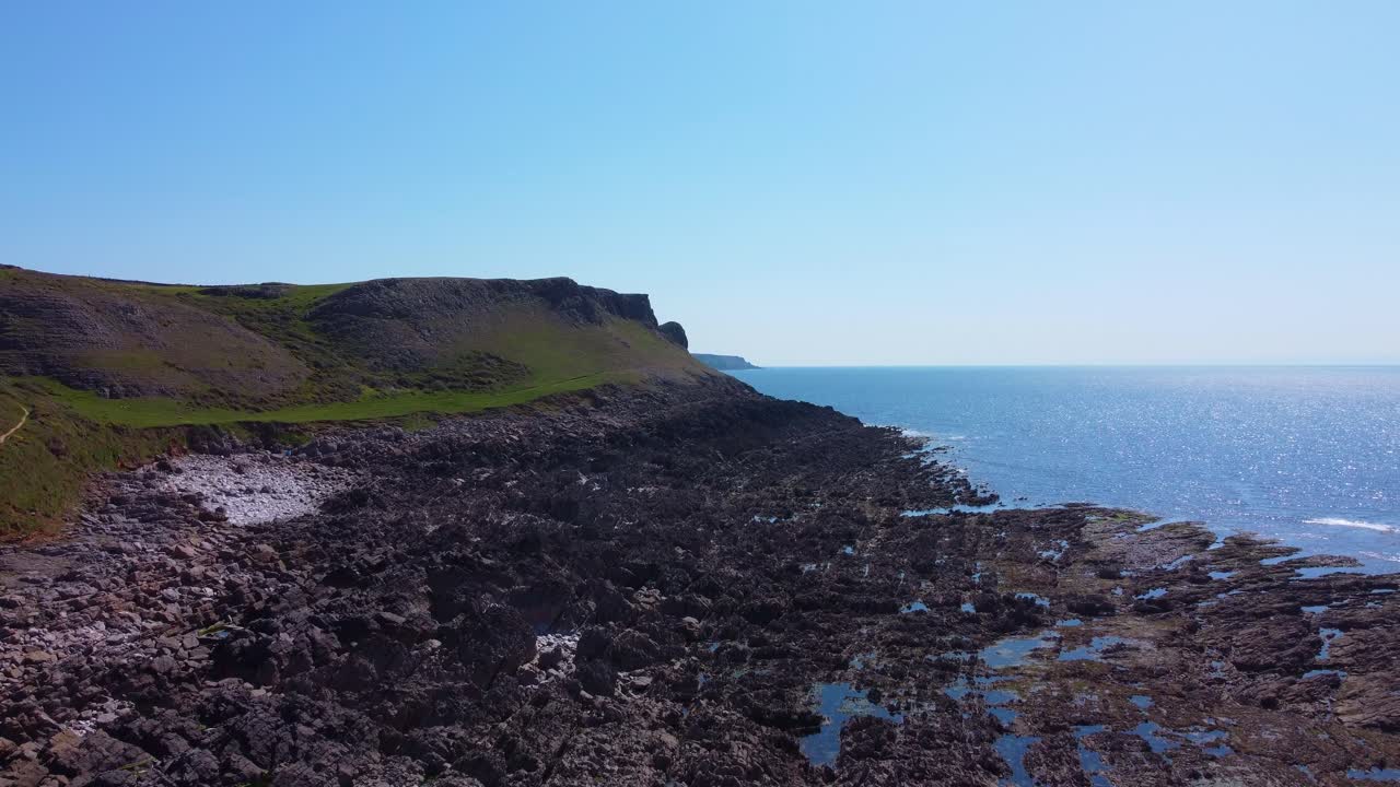 Slow Rising Drone Aerial View of Steep Cliff Tops with Bay at Low Tide and Sun Reflecting on Sea Water Creating Wide Picturesque View.