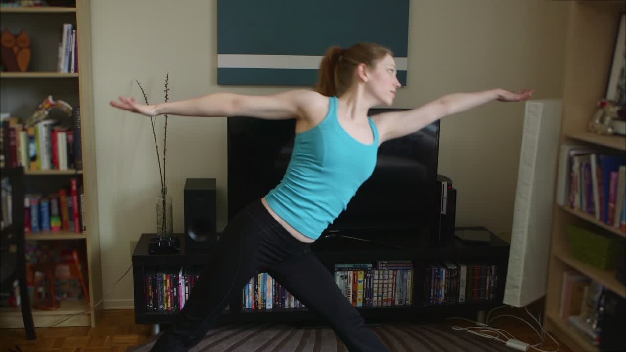 A young, fit woman, dressed in a blue tank top and black yoga pants, doing yoga in her apartment.