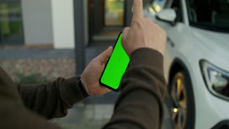 persona sosteniendo un teléfono inteligente con pantalla verde, probablemente para grabación de video o fines tutoriales, frente a una casa y un automóvil.