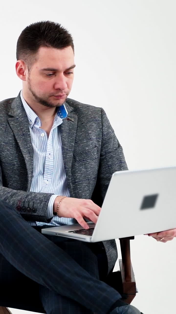 Portrait of serious businessman with a laptop. Thoughtful young man sitting in a chair and working a laptop isolated on white background. Vertical video