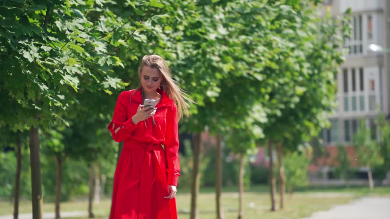 Lovely model with a cellphone. Beautiful young woman in bright red dress with a phone on the green trees background in summer. Slow motion.