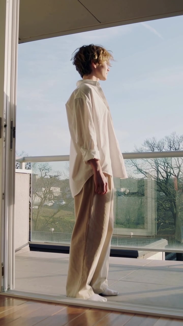 Video verticale: Uomo in piedi con camicia bianca che guarda il balcone dalla porta a vetri, con la luce del sole che crea giochi di luce.