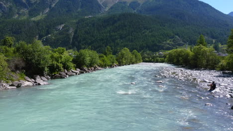 turquoise-blue-river-surrounded-by-lush-green-trees-in-the-mountains-of-Zernez-Switzerland-on-summer-day,-aerial