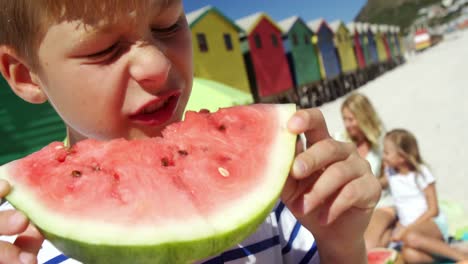 Junge-Isst-Wassermelone,-Während-Die-Familie-Im-Hintergrund-Am-Strand-Sitzt