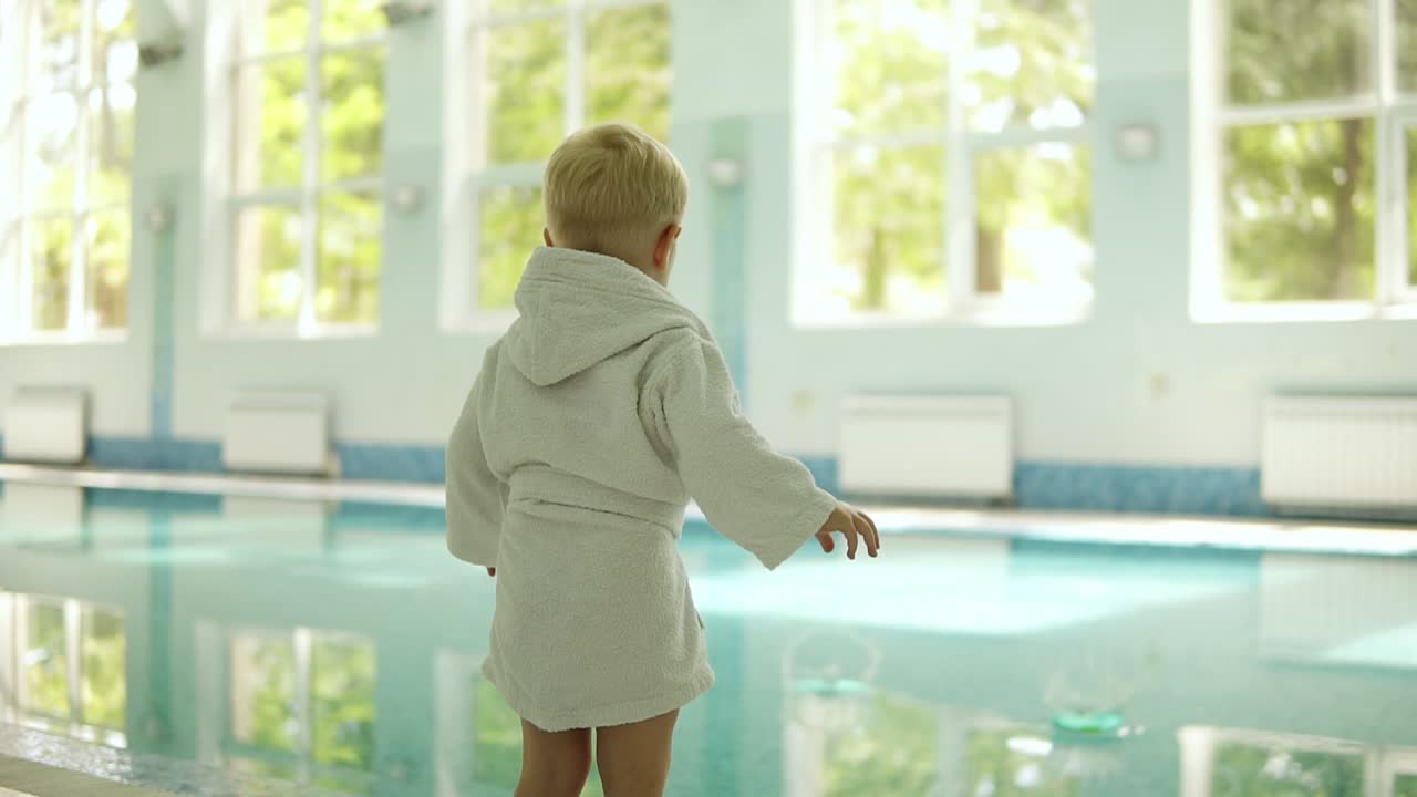 Backside view of a little boy in bathrobe throwing balls from both hands into the swimming pool. Playful. Outside the pool. Pure water