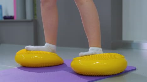 close-up of young child balancing on yellow rubber squat pads while kneeling on a purple mat, child wears white socks and pink shorts. background features gym equipment