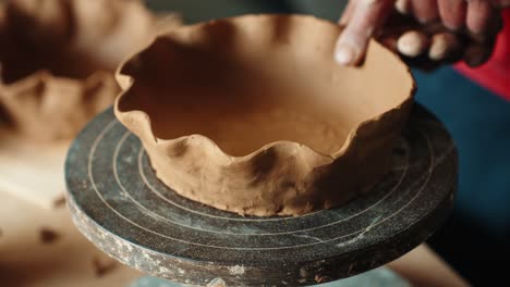 a close-up shot of a ceramic artist's hands meticulously shaping and creating a decorative wavy rim on a rustic, handmade clay bowl resting on a banding wheel in a studio