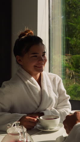 couple talks drinking tea by window. young people in bathrobes enjoy weekend drinking hot beverage in riverside hotel. concept of vacation relaxation