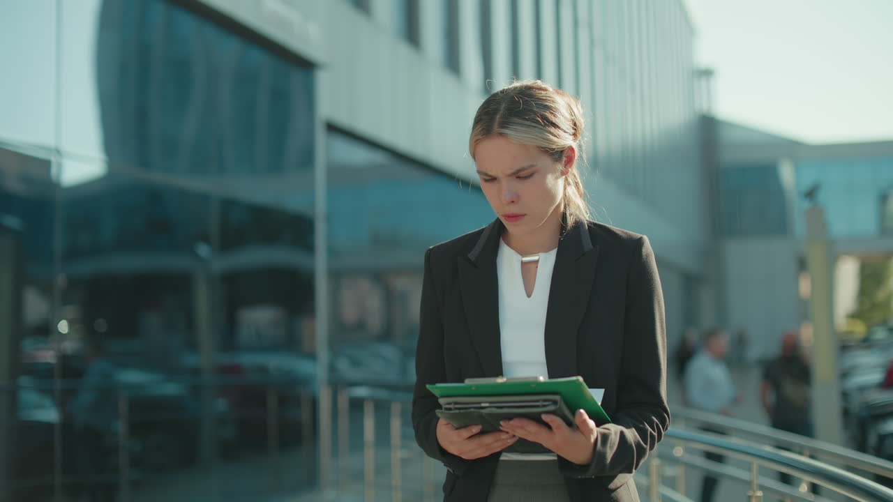 Focused female CEO outdoors in formal attire reviewing documents in folder while walking near modern glass building with pedestrians and parked cars in background on bright sunny day