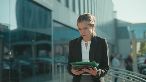 focused female ceo outdoors in formal attire reviewing documents in folder while walking near modern glass building with pedestrians and parked cars in background on bright sunny day