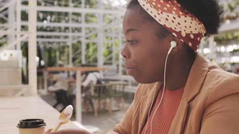 Mujer-Comiendo-Helado-Sentada-En-La-Terraza-Del-Bar-Mientras-Habla-Por-Teléfono-Con-Auriculares