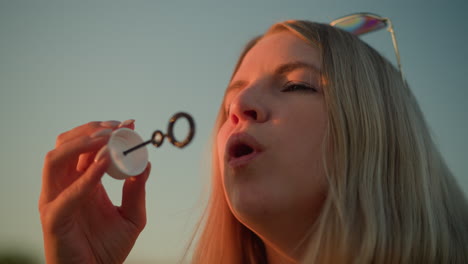 primer plano de una mujer blanca con gafas de sol en la cabeza soplando burbujas al aire libre durante la puesta del sol, la luz del sol se refleja cálidamente en su cara, iluminando sus rasgos mientras sostiene la varita de burbujas