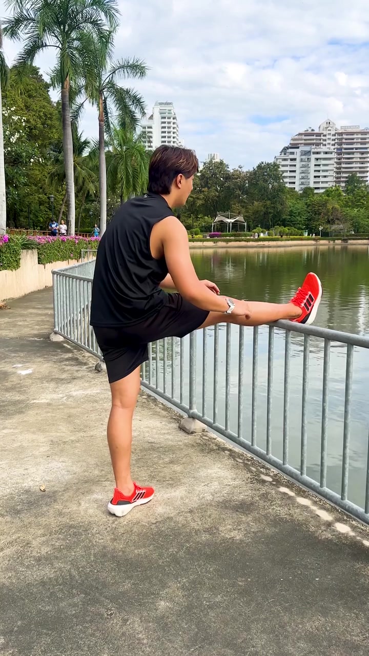 Young man performs dynamic leg stretches beside lakeside railing in sunny urban park setting