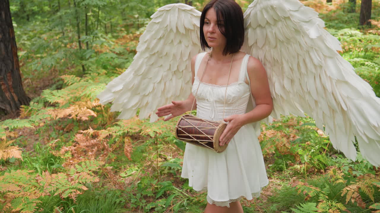 Attrice cinematografica vestita come una divinità con grandi ali bianche che cammina lentamente attraverso la foresta, suonando delicatamente un tamburo kalimba con un ritmo pacifico, circondata dalla luce del sole e dalle felci