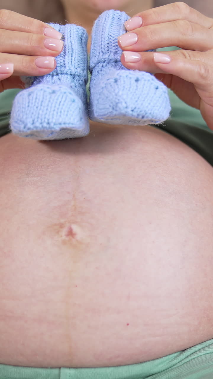 Bare stomach of a future mother. Pregnant woman playing with little knitted socks on her belly lovingly. Close-up. Vertical video