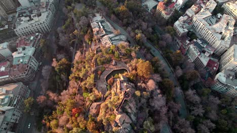 aerial rising over top of santa lucia hill covered with autumnal trees and pedro de valdivia square between santiago buildings, chile