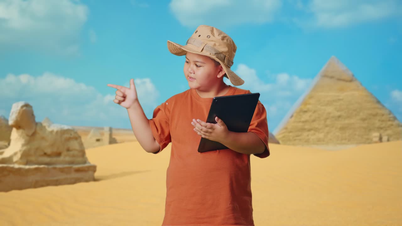 Asian Boy With A Hat Using A Tablet Then Smiling And Pointing To Side While Traveling In Giza Pyramid. Boy Researcher Examines Something, Travel Tourism Adventure Concept