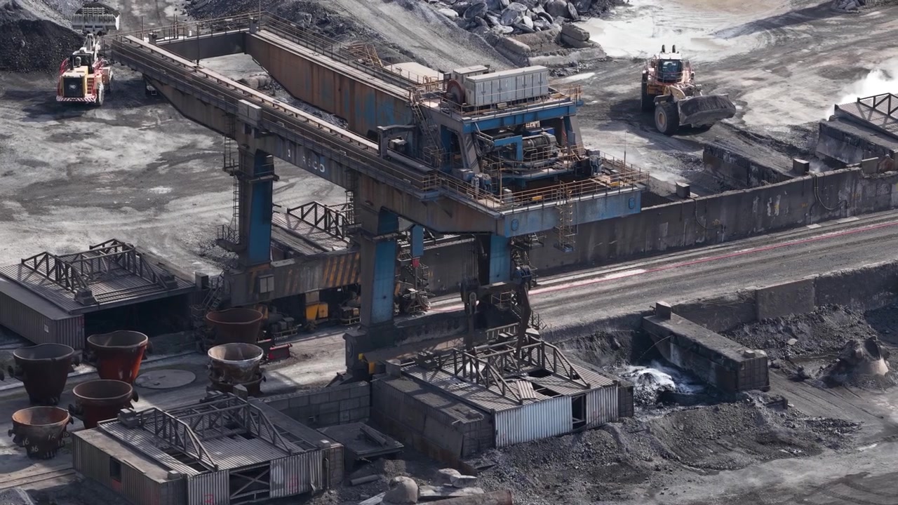 Industrial machinery operating in a large steel production facility, with a bulldozer moving piles of coal and ore near a gantry crane and other heavy equipment in the plant yard