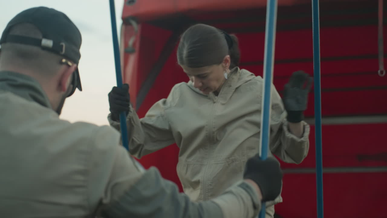 close up of man and woman working together to insert long metal poles into hot air balloon basket by red trailer on grassy field, focus on coordinated effort and safety setup before flight