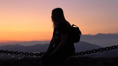 Junge-Frau-Sitzt-Auf-Einem-Schwingenden-Kettengeländer-Vor-Einem-Wunderschönen-Rosa-orange-gelben-Berghintergrund-Mit-Blick-Auf-Den-Sonnenuntergang-In-Kalifornien-Und-Neigt-Sich-Nach-Oben