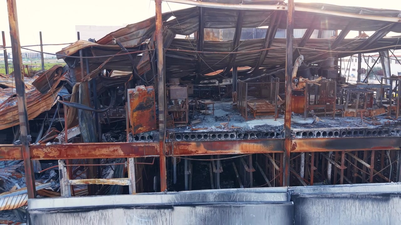 Burned-out industrial building with charred steel framework, twisted metal roofing, rusted structural beams, and ash-covered debris scattered across the destroyed interior after a fire