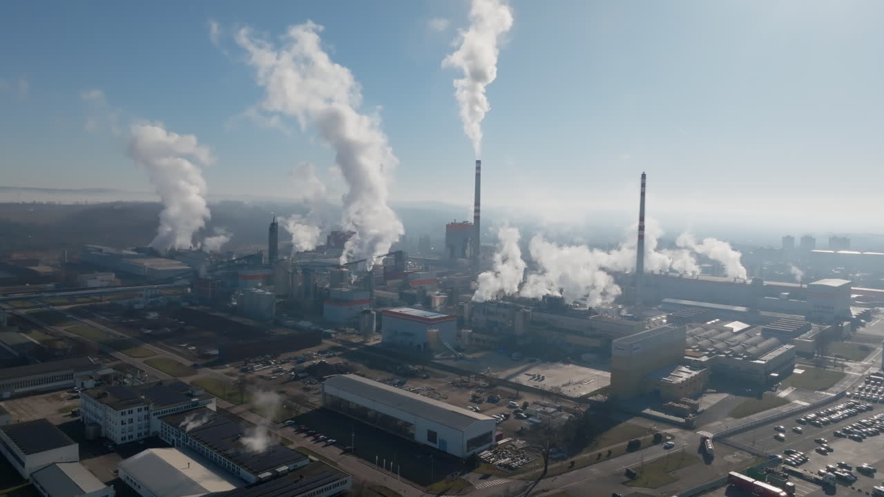 Ripresa con drone lungo l'area industriale fluviale a Štětí, con ciminiere che emettono vapore e riflessi specchiati sull'acqua calma sotto un sole splendente