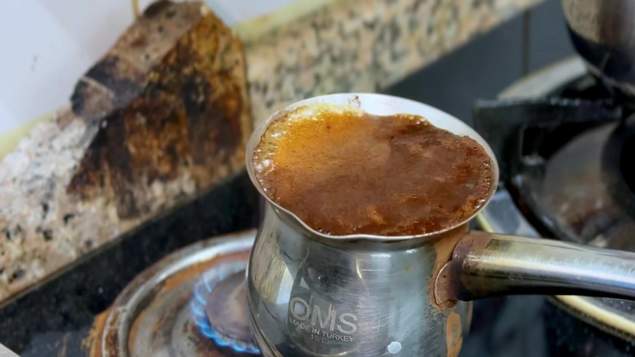 A close-up view of dark Turkish coffee boiling and frothing inside an OMS metal pot. The traditional beverage brews over a blue gas stove flame in an indoor kitchen setting during a morning routine.
