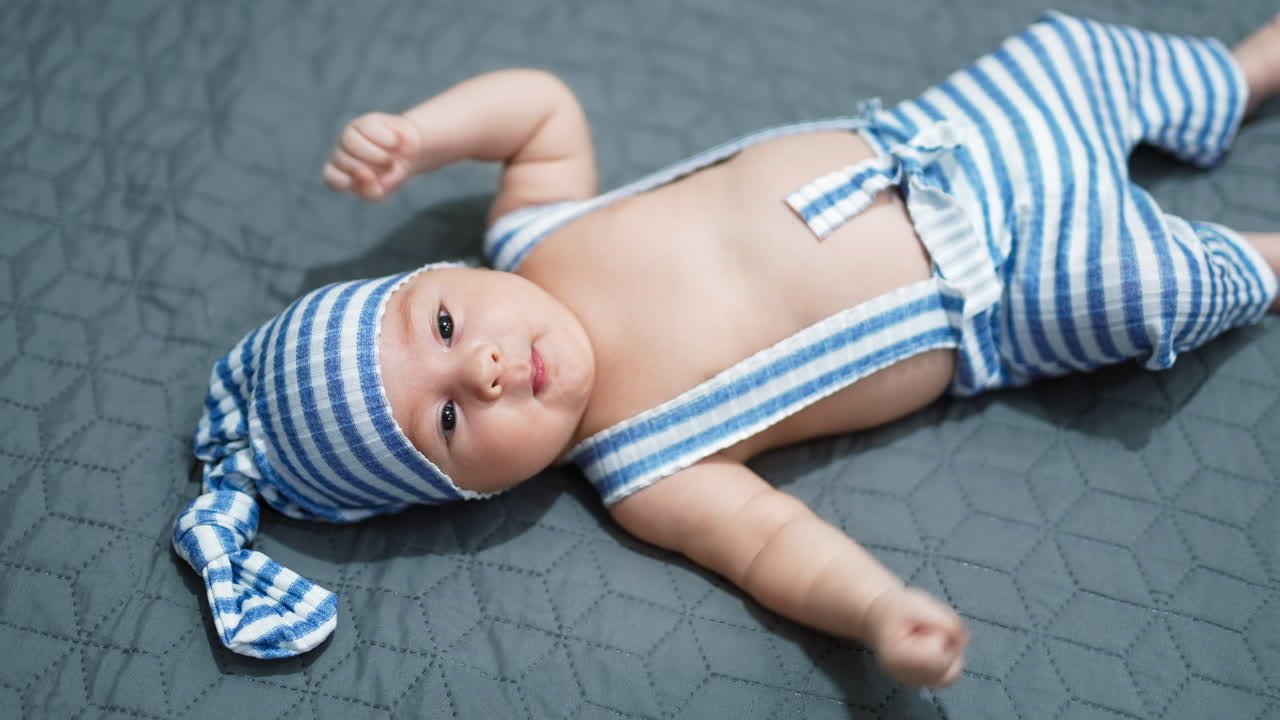 Baby boy wearing striped costume lying on the bed. Baby in a cap on the grey background. View from above.