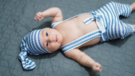 baby boy wearing striped costume lying on the bed. baby in a cap on the grey background. view from above.