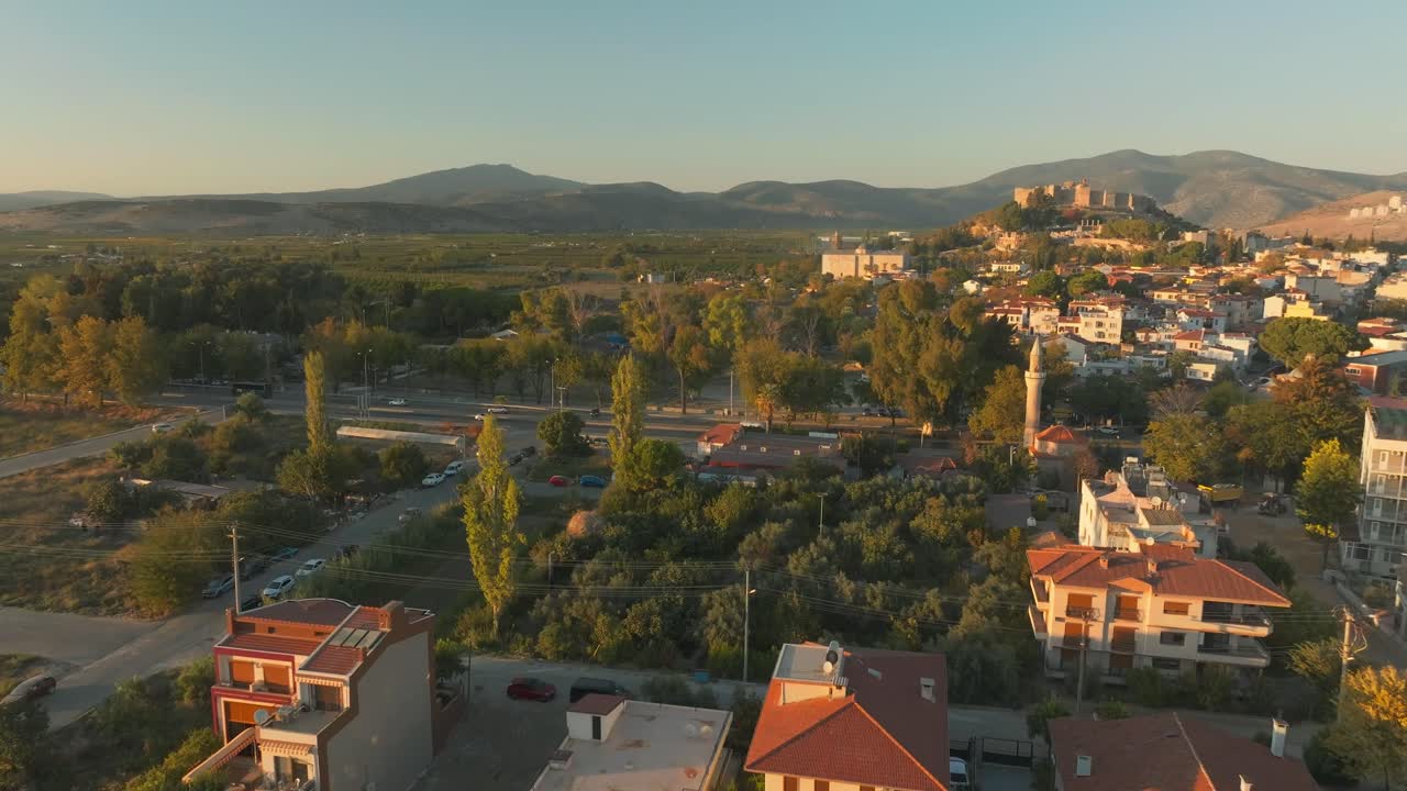 Drone glides over treetops, unveiling Selçuk Castle, distant mountains, at golden hour.