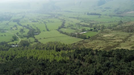 Drone-shot-panning-down-over-trees-and-farmland-with-the-village-Castleton-in-the-background,-Peak-District