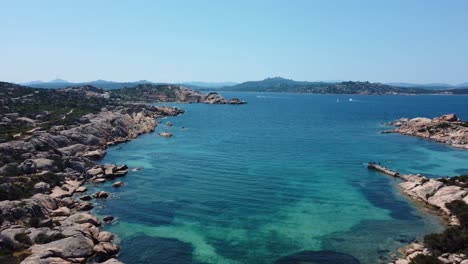 flying along an idyllic natural coast beach rock bay at la maddalena on the tourist vacation island sardinia in italy with sun, clear blue turquoise and calm water