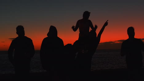 Silueta-Oscura-Que-Muestra-A-Un-Grupo-De-Jóvenes-Celebrando-Al-Aire-Libre-Y-Disfrutando-De-La-Puesta-De-Sol-De-Color-Rojo-En-El-Fondo