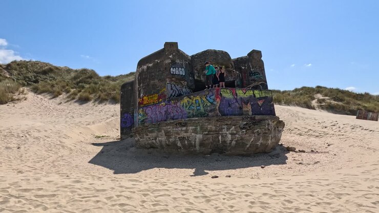 Una telecamera fissa cattura un bunker della Seconda Guerra Mondiale illuminato dal sole e coperto di graffiti su una spiaggia sabbiosa a Dunkerque, Francia, con cieli azzurri e sereni sopra.