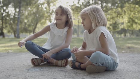 two caucasian girls sitting cross-legged and drawing with chalks on asphalt