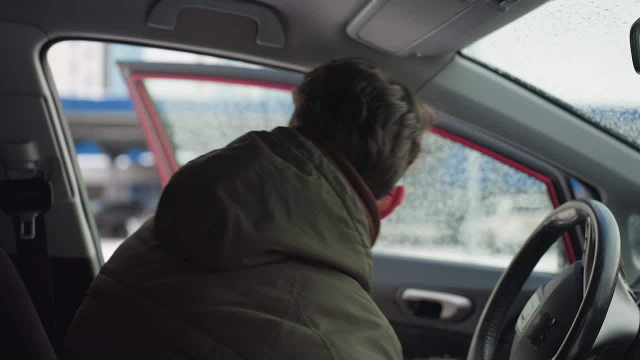 Close up of man in winter jacket stepping out from car during cold weather with water droplets on window, urban setting and passing vehicle visible through blurred frosted glass