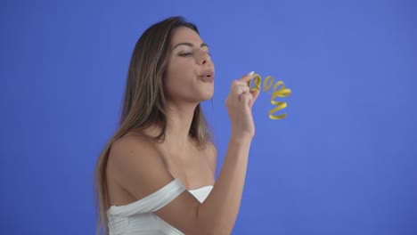 beautiful girl blows a yellow party streamer, isolated on blue background.