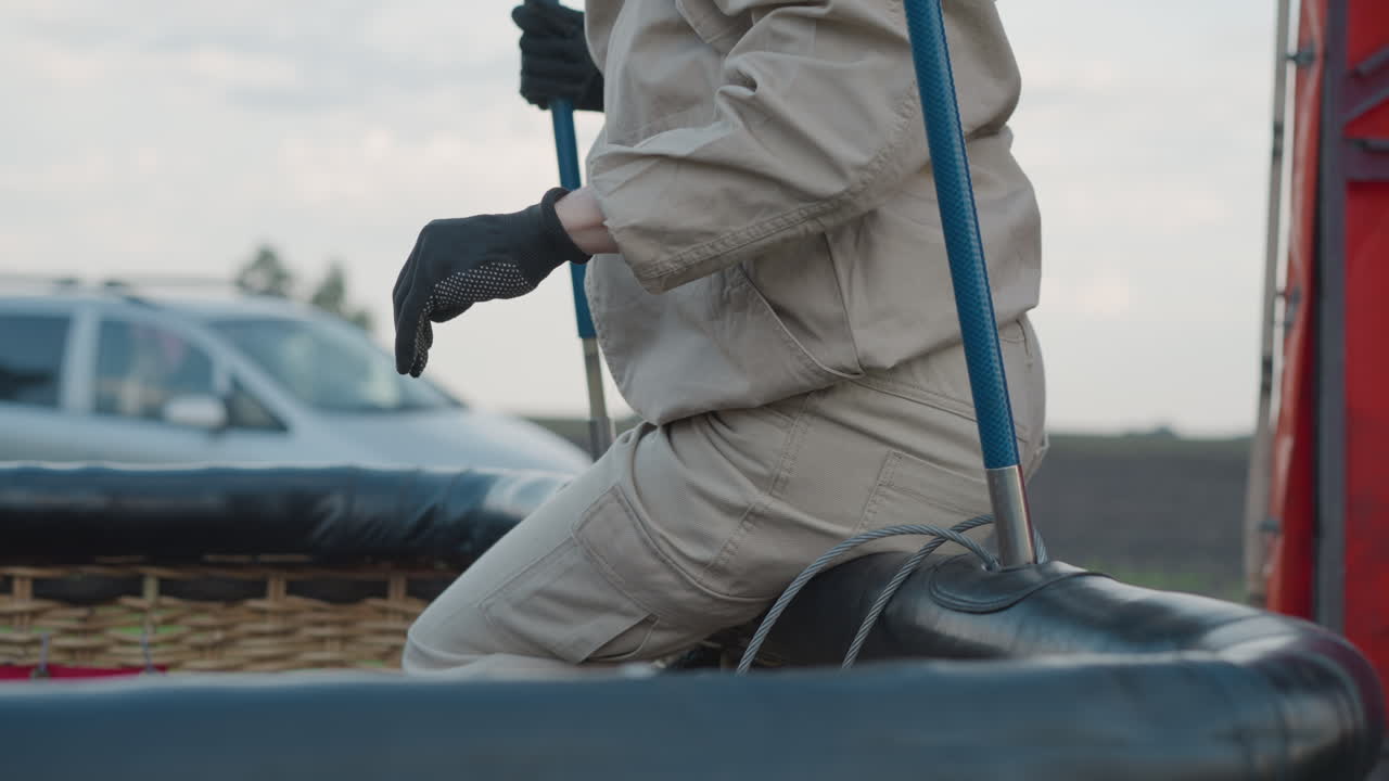 person seated on hot air balloon basket adjusting support pole by roadside with parked vehicle visible background wearing protective gloves under cloudy sky