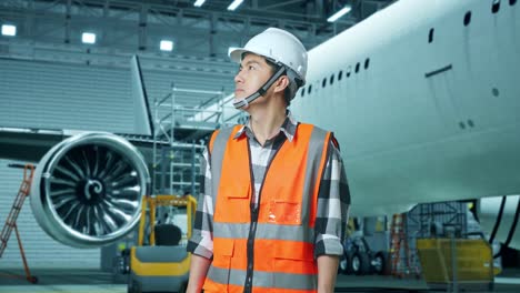 ingeniero asiático con casco de seguridad de pie con aviones en el hangar, mirando a su alrededor, comprobando