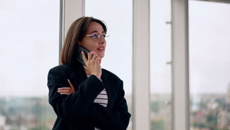 caucasian brunette having phone conversation indoors. portrait of a positive lady in glasses speaking on the smartphone near the window.