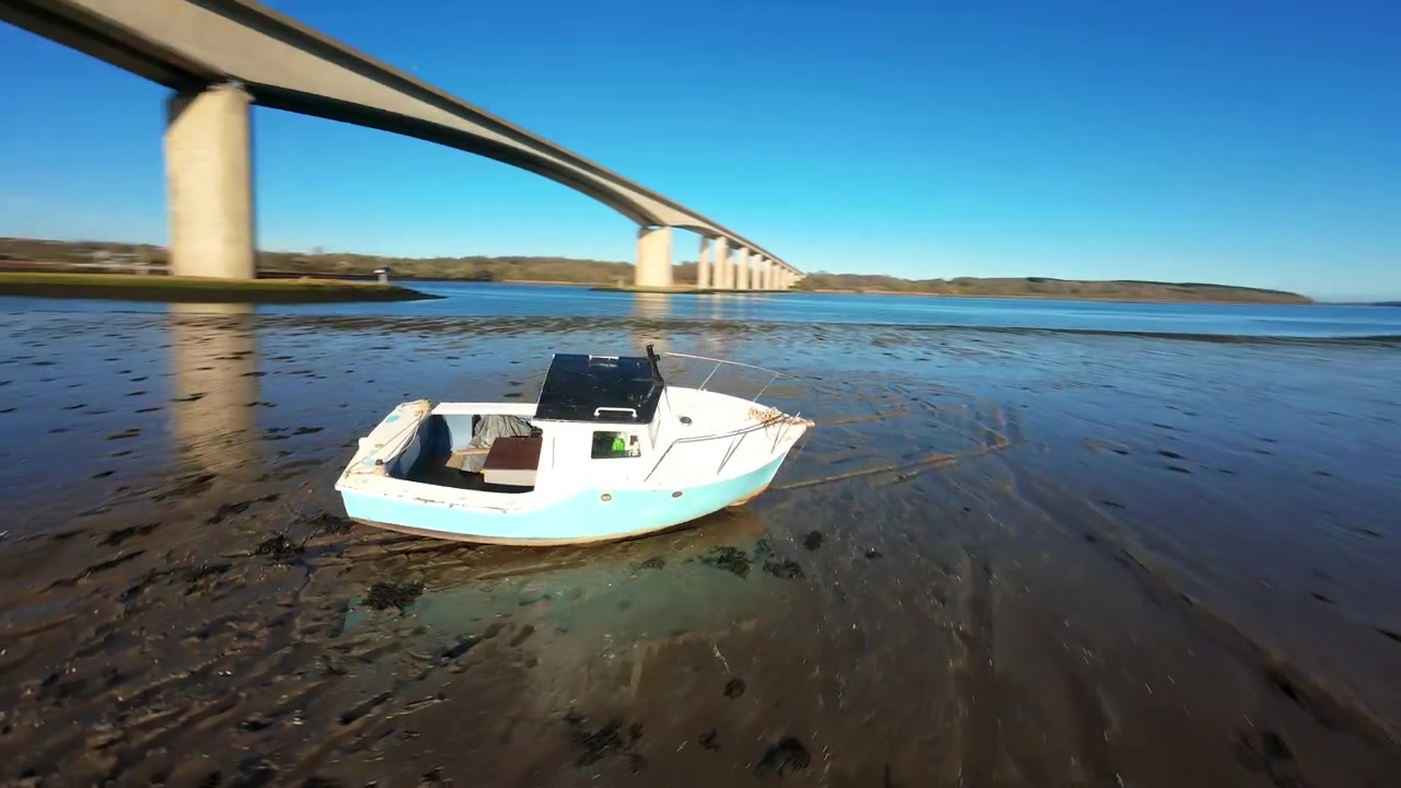 Kumlu sahilde terk edilmiş bir balıkçı teknesinin etrafında alçak uçuş yapan dinamik FPV Drone.