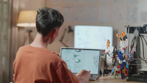 vista trasera de un niño con impresión 3d pensando mientras diseña una mano cyborg en una computadora portátil en casa